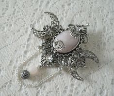 Rose Quartz Triple Moon Brooch This beautiful silver plated brooch has silver plated crescent moons, silver plated embellishments, a rose quartz setting, rhinestone, metal chain, silver plated filigree bead and rose quartz bead. Can also be worn as a cloak pin. It is 2.75 inches wide and 3.75 inches long with bead drop and chain. Gothic Brooch Jewelry Gift, Spiritual Silver Brooch, Spiritual Silver Jewelry Brooch, Spiritual Silver Brooch For Gift, Silver Bohemian Brooch Jewelry, Silver Bohemian Jewelry Brooch, Handmade Silver Bohemian Brooch, Cloak Pin, Embossed Leather Journal