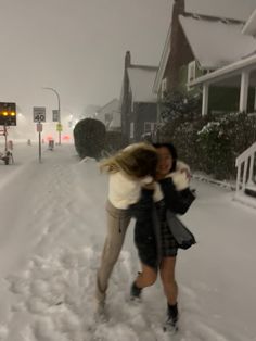 two people are hugging in the snow on a street with houses and cars behind them