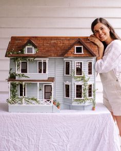 a woman standing next to a doll house on top of a white cloth covered table