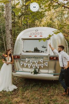 a bride and groom standing next to an airstream in the woods with their hands up