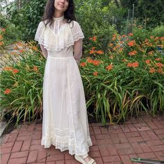 a woman in a white dress standing next to some bushes and flowers with her hands on her hips