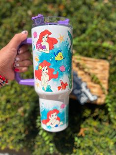 someone holding up a cup with ariel the little mermaid on it in front of some bushes