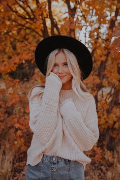 a woman wearing a black hat and sweater posing for a photo in front of trees