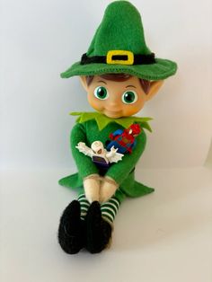 a green elf doll sitting on top of a white surface with his legs crossed and eyes wide open