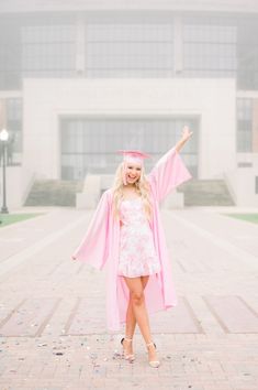 a woman in a pink graduation gown and cap is standing on the sidewalk with her arms outstretched