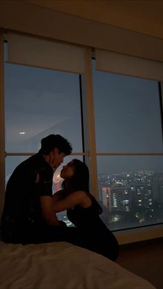 a man and woman are kissing in front of large windows at night with the city lights lit up behind them