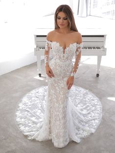 a woman standing in front of a piano wearing a wedding dress with long sleeves and an off the shoulder design