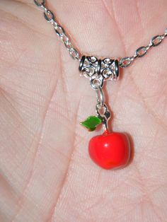 a hand holding a small red apple charm on a silver plated chain with a green leaf attached to it