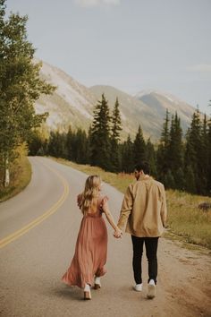 a man and woman holding hands walking down the road with mountains in the back ground