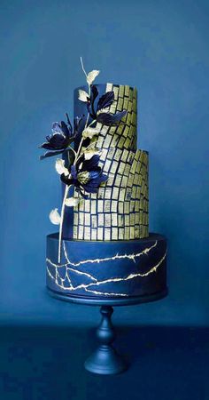 a three tiered blue and gold wedding cake with flowers on the top, against a blue background