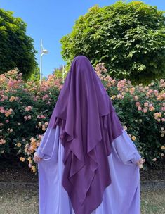 a woman wearing a purple veil standing in front of some bushes and flowers with her back turned to the camera