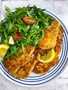 a white and blue plate topped with fish, salad and lemon wedges on top of a marble table