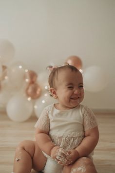 a smiling baby sitting on the floor in front of balloons