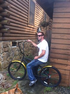 a man sitting on a bike next to a brown dog and a log cabin building