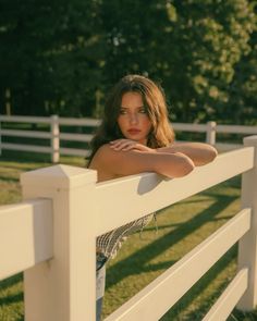 a beautiful young woman leaning on a white fence in the grass with her arms crossed