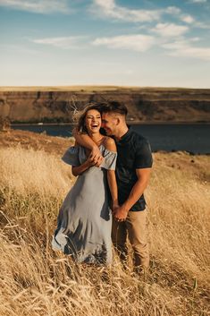 a man and woman standing in tall grass by the water with their arms around each other