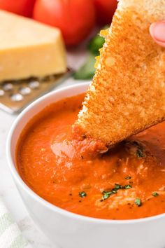 a hand dipping some bread into a bowl of tomato soup with cheese on the side