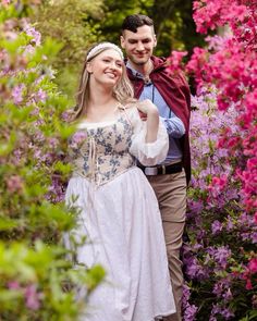 a young man and woman dressed in period costumes walking through flowers with their arms around each other