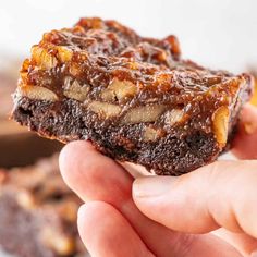 a hand holding up a piece of brownie with nuts on top and chocolate in the background