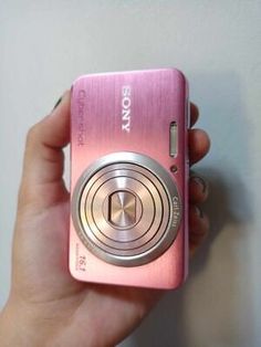 a hand holding a pink camera in front of a white wall with the word sony on it