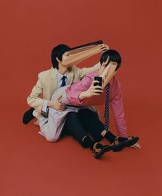 two people sitting on the ground with one person covering his eyes and holding a cell phone