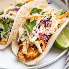 three fish tacos with cole slaw and lime wedges on a white plate