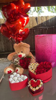 a teddy bear sitting on top of a table next to heart shaped balloons and flowers