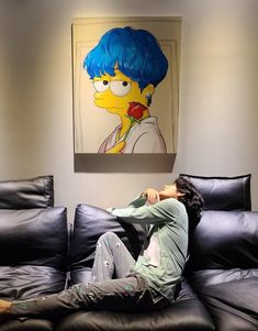 a man sitting on top of a black couch next to a simpsons painting in a living room