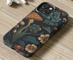 an iphone case with flowers and mushrooms on it, sitting on a wooden table next to ear buds