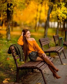 a beautiful young woman sitting on top of a wooden bench in a park next to trees