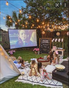 kids are sitting on the grass in front of an outdoor movie screen with lights strung over them