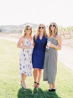 three women standing next to each other on top of a grass covered field wearing sunglasses