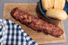 a wooden cutting board topped with meat next to buns and a bowl of bread