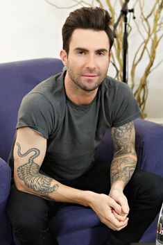 a man sitting on a blue couch with tattoos on his arm and chest, smiling at the camera