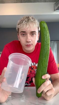 a young man holding up a large cucumber next to a plastic cup