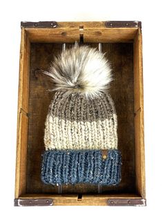 a knitted hat with a fur pom sits in a wooden box on display