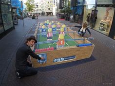 a man kneeling down next to a giant game on the side of a street with people walking by