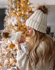 a woman holding a baby in front of a christmas tree wearing a white sweater and hat