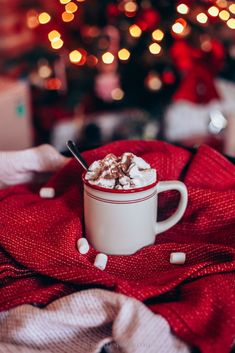 a white cup filled with hot chocolate and marshmallows on top of a red blanket