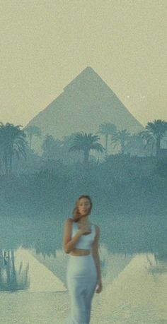 a woman standing in front of a pyramid with palm trees behind her and the sky reflecting on the water