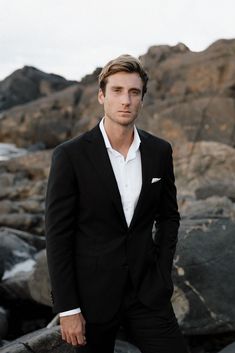 a man in a black suit and white shirt standing on rocks near the ocean with his hands in his pockets