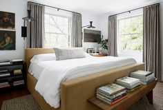 a bed with white sheets and pillows in a bedroom next to two windows filled with books