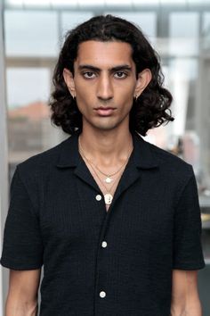 a young man with long hair wearing a black shirt and silver necklace standing in front of a window