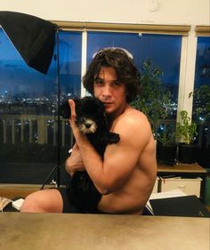 a shirtless man holding a small dog in front of a window with the city lights behind him