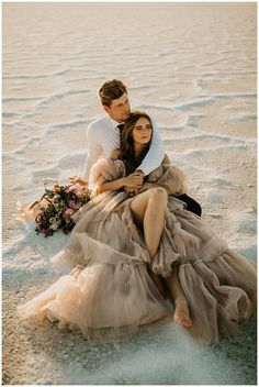 a man and woman are sitting on the sand in their wedding gowns, holding each other