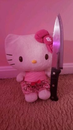 a hello kitty stuffed animal sitting next to a large knife on the floor in front of a pink wall