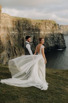 a bride and groom standing on the cliff by the ocean with their wedding dress blowing in the wind