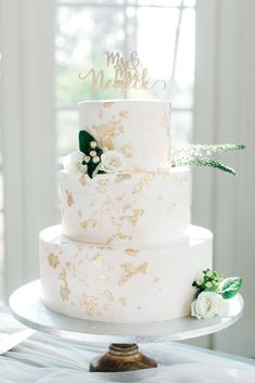 a white and gold wedding cake sitting on top of a metal stand with greenery