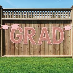 a graduation banner that says grad in pink letters on a wooden fence with grass