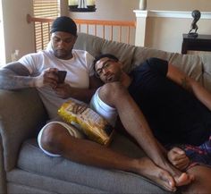 two men sitting on a couch looking at their cell phones while one man holds a bag of chips